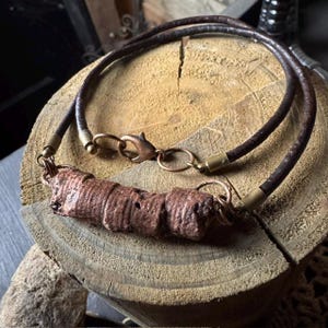 May include: A handcrafted necklace featuring a dark brown leather cord and a textured, reddish-brown central pendant. The necklace has copper-colored clasps and connectors. The pendant has a unique, organic shape, adding a rustic touch to the jewelry.
