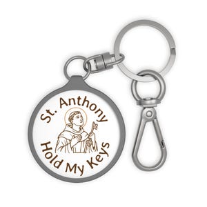 May include: A round keychain with a gray border and a white center featuring an illustration of St. Anthony holding a key. The text "St. Anthony Hold My Keys" is printed around the image. The keychain has a silver clasp and key ring.