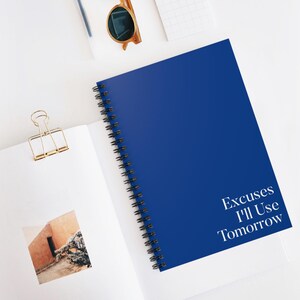 Puede incluir: Un cuaderno azul cobalto con espiral con el texto "Excuses I'll Use Tomorrow" en blanco. El cuaderno está abierto, revelando una página blanca con un clip dorado y una pequeña foto. Gafas de sol y un bolígrafo también son visibles.