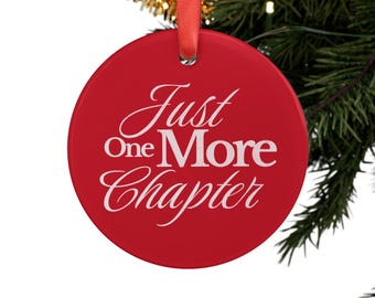 Puede incluir: Un adorno navideño redondo y rojo con el texto blanco "Just One More Chapter". El adorno cuelga de una cinta roja sobre un fondo de árbol de Navidad.