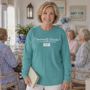 May include: A teal long-sleeve shirt featuring the white text "Emotionally Attached TO AN IMPOSSIBLE HAND" above three small playing card icons. The shirt has a crew neckline and a relaxed fit, made of a solid-coloured fabric with a smooth texture.