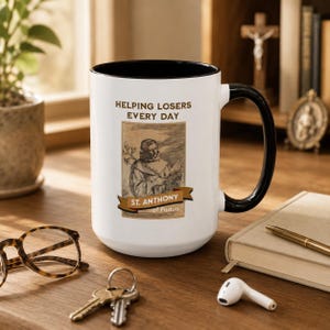 May include: A white ceramic mug with a black handle and interior rim. The front features a brown-toned illustration of Saint Anthony of Padua with the text "HELPING LOSERS EVERY DAY" above and a banner reading "ST. ANTHONY of Padua" below.