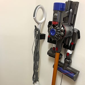 May include: A white and gray steam mop and a black and orange cordless vacuum cleaner are mounted on a wall with a black and white wall mount. The vacuum cleaner has a blue and red dustbin and a blue and red power button.