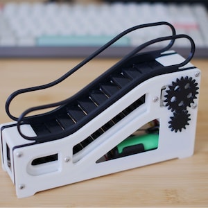 May include: A white plastic model of an escalator with a black rubber handrail and black gears. The model is on a wooden surface.