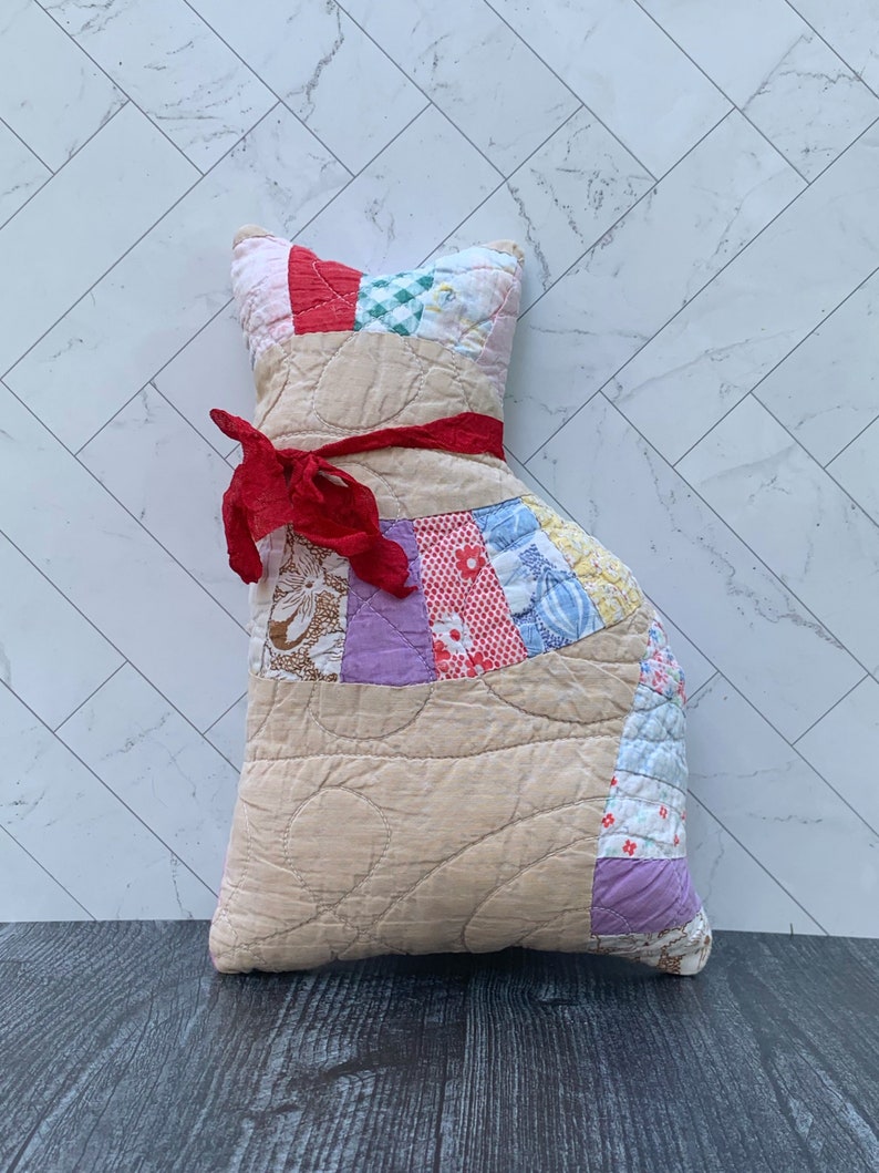 May include: A handmade cat-shaped pillow made from a patchwork quilt. The pillow features a tan body with a red ribbon tied around the neck. The head and back are made of colorful fabric squares. The pillow is approximately 15 inches tall.