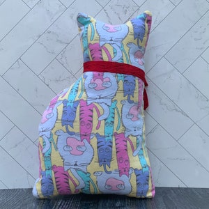 May include: A cat-shaped pillow with a colorful pattern of cartoon cats in various poses. The pillow features a yellow background and is adorned with a red ribbon around its middle. The cat illustrations are in shades of pink, blue, and purple.