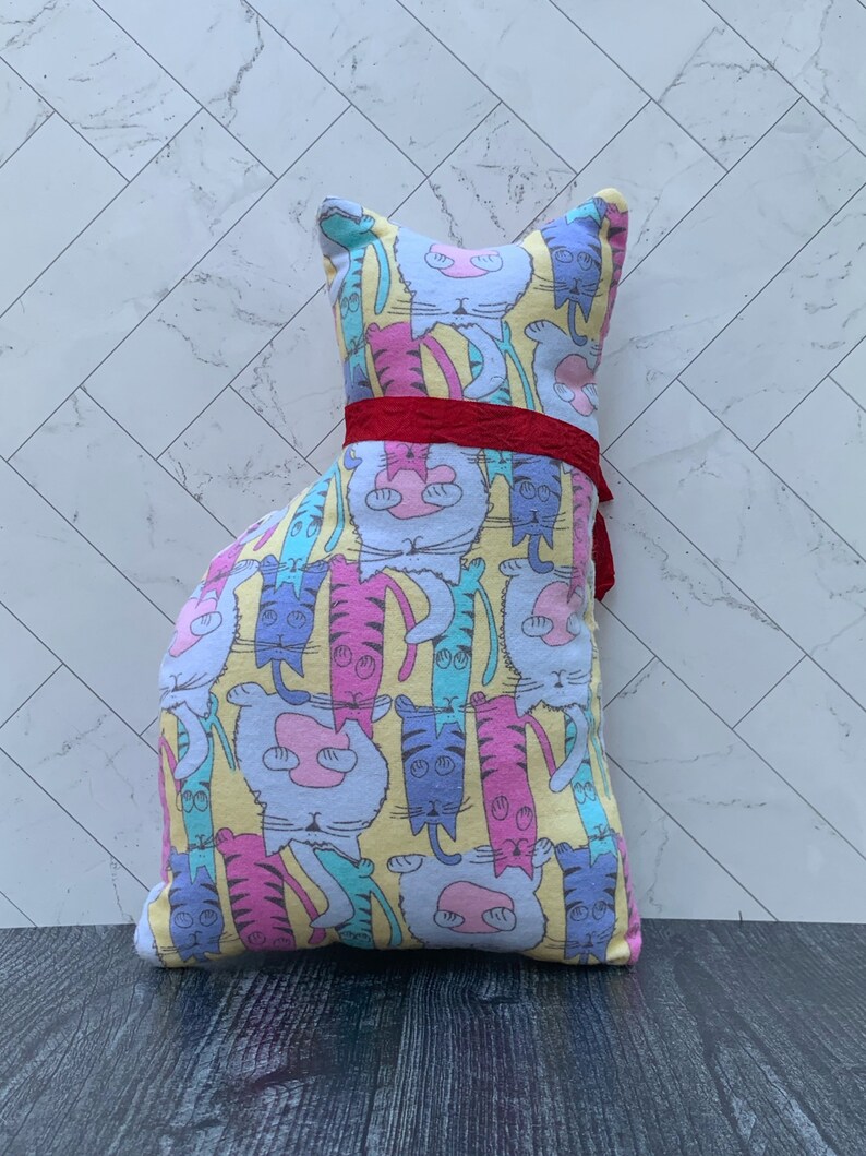 May include: A cat-shaped pillow with a colorful pattern of cartoon cats in various poses. The pillow features a yellow background and is adorned with a red ribbon around its middle. The cat illustrations are in shades of pink, blue, and purple.