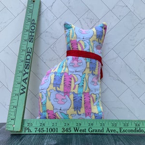 May include: A cat-shaped pillow with a colorful cat pattern on a yellow background, accented with a red ribbon. The pillow is approximately 10 inches tall. A green ruler with the text "WAYSIDE SHOP" and phone number is in the image.