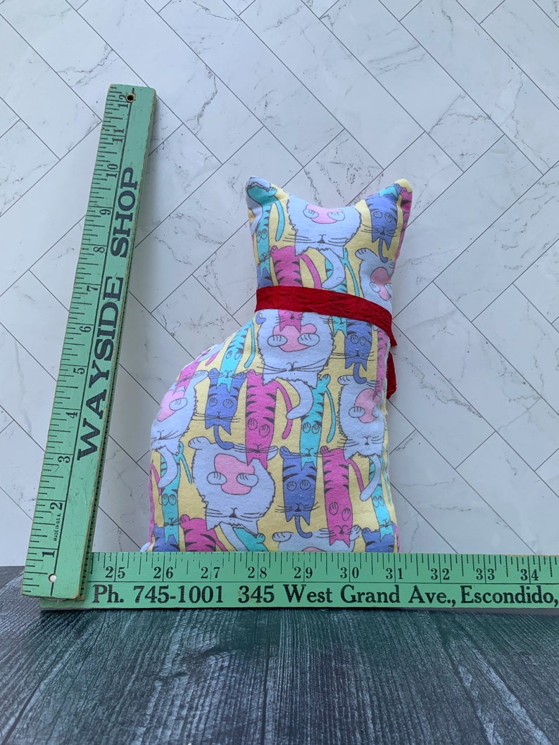 May include: A cat-shaped pillow with a colorful cat pattern on a yellow background, accented with a red ribbon. The pillow is approximately 10 inches tall. A green ruler with the text "WAYSIDE SHOP" and phone number is in the image.