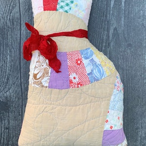 May include: A handmade cat-shaped pillow with a patchwork design. The pillow features a tan body with quilted stitching and a head made of colorful fabric scraps. A red ribbon is tied around the neck. This decorative pillow is a unique home decor item.