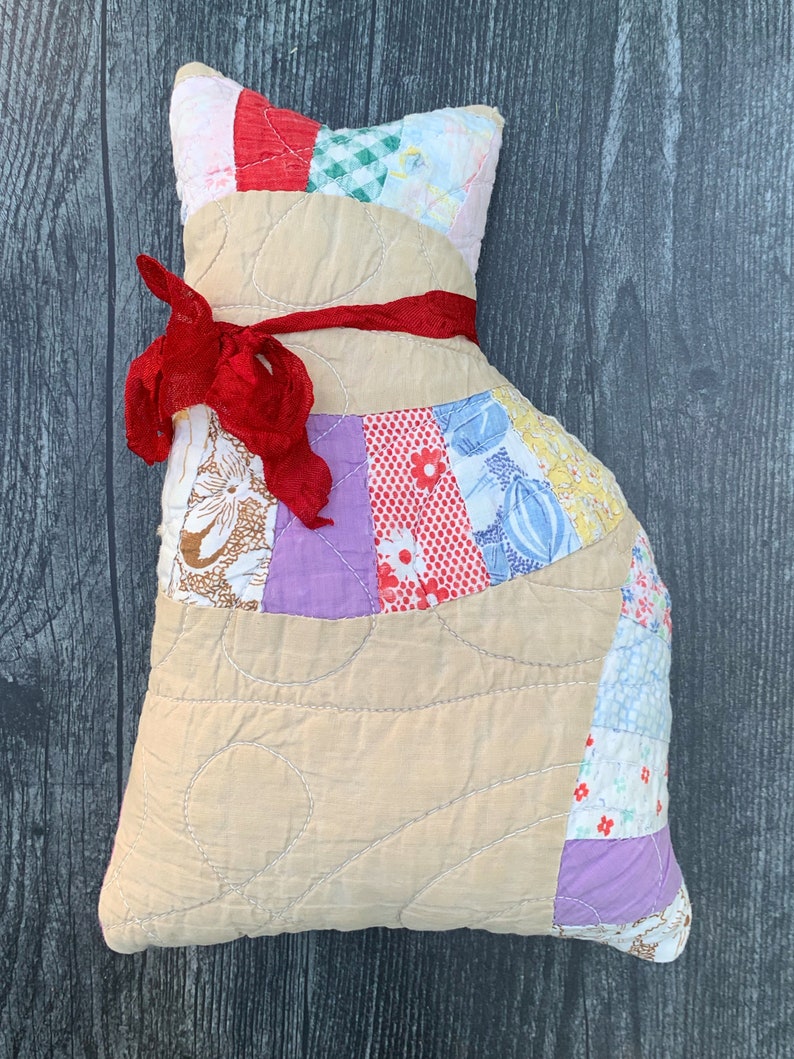 May include: A handmade cat-shaped pillow with a patchwork design. The pillow features a tan body with quilted stitching and a head made of colorful fabric scraps. A red ribbon is tied around the neck. This decorative pillow is a unique home decor item.