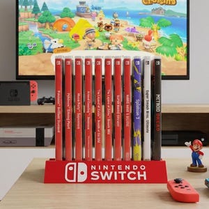 Può includere: Un supporto rosso per dodici custodie di giochi Nintendo Switch, posizionato su un tavolo di legno. Il supporto presenta il logo bianco Nintendo Switch. Nelle vicinanze si trovano due controller, tre piccole statuette e una ciotola di popcorn.