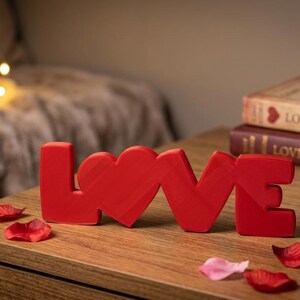 Può includere: Una scritta decorativa rosso brillante che forma la parola LOVE, in cui la lettera O è sostituita da una forma a cuore. L'oggetto è posizionato su una superficie in legno accanto a un calice di vino rosso, due libri e petali sparsi.
