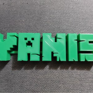 Può includere: Un cartello con nome stampato in 3D di colore verde che dice "YANIS" con un viso di creeper pixelato nella "A".