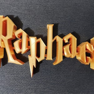 Può includere: Un'etichetta nominativa in 3D stampata in oro con il nome "Raphael" in un carattere stilizzato.