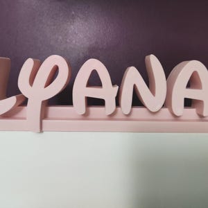 Könnte beinhalten: Ein hellrosa Holzschild mit dem Namen "LYANA". Die Buchstaben sind in einer abgerundeten Schriftart gehalten und an einer passenden Basis befestigt. Das Schild wird vor einer lila Wand und einer weißen Oberfläche präsentiert.
