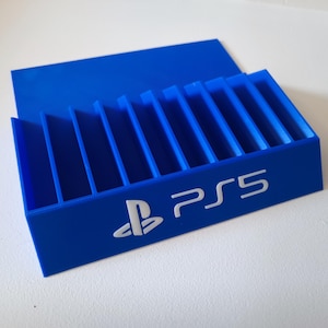 Op de afbeelding: Een blauwe PS5-spelhouder met meerdere sleuven voor spelopslag. De houder heeft het PS5-logo en het PlayStation-symbool in het wit. De houder is gemaakt van een solide, duurzaam materiaal.