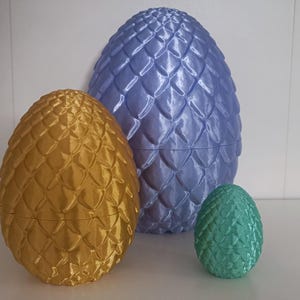 Puede incluir: Tres adornos con forma de huevo de dragón, uno azul, uno dorado y uno verde. Los adornos están hechos de un material texturizado que se asemeja a escamas de dragón.