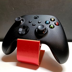 Op de afbeelding: Zwarte Xbox-controller op een rode standaard. De controller heeft gekleurde knoppen en twee joysticks. De standaard is gemaakt van een gevouwen, glanzend rood materiaal.