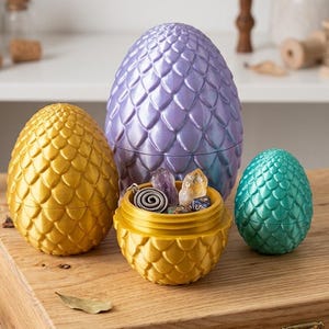 Peut inclure: Trois œufs de style dragon avec un motif d'écailles texturé. Un grand œuf est violet métallisé, un œuf moyen est doré métallisé et un petit œuf est bleu sarcelle métallisé. L'œuf doré est ouvert, révélant des cristaux et une breloque en métal.