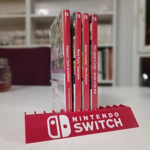 Puede incluir: Un soporte rojo para juegos de Nintendo Switch con el logotipo de Nintendo Switch en blanco. El soporte muestra cuatro cajas de juegos: "Pokémon - Perle Scintillante", "Mario Party Superstars", "Animal Crossing - New Horizons" y "The Legend of Zelda: Breath of the Wild".