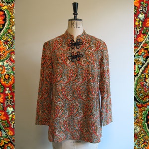 May include: A long-sleeved, orange, green and black paisley patterned shirt with a mandarin collar and black button fastenings. The shirt is on a white mannequin.
