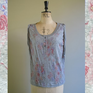 Pode incluir: Uma camisa cinza de manga curta com estampa floral em tons de rosa, azul e amarelo. A camisa tem um fecho de botões na frente e um caimento descontraído.