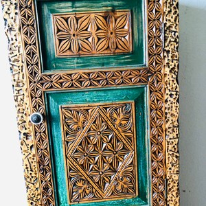 K&ouml;nnte beinhalten: Ein hoher Holzschrank mit einer geschnitzten, gr&uuml;nen T&uuml;r. Die T&uuml;r hat komplizierte Blumenmuster und einen goldfarbenen Rahmen.