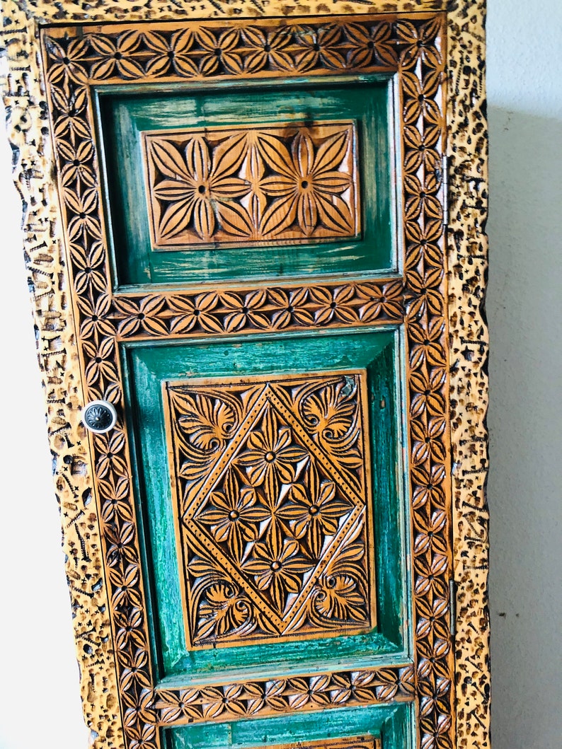 K&ouml;nnte beinhalten: Ein hoher Holzschrank mit einer geschnitzten, gr&uuml;nen T&uuml;r. Die T&uuml;r hat komplizierte Blumenmuster und einen goldfarbenen Rahmen.