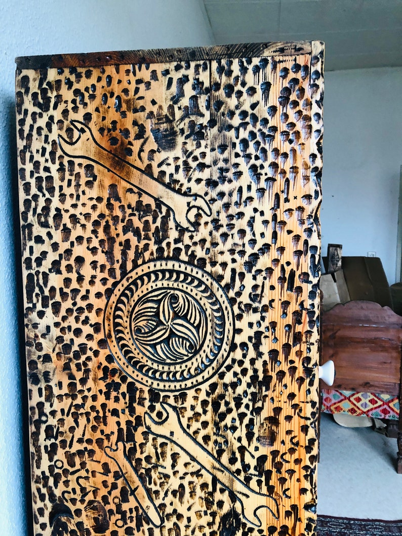 K&ouml;nnte beinhalten: Ein Holzschrank mit rustikalem, abgenutztem Finish. Die Schrankt&uuml;r zeigt ein geschnitztes Design mit zwei Schraubenschl&uuml;sseln und einem kreisf&ouml;rmigen Muster. Das Holz ist hellbraun mit dunkelbraunen Akzenten.