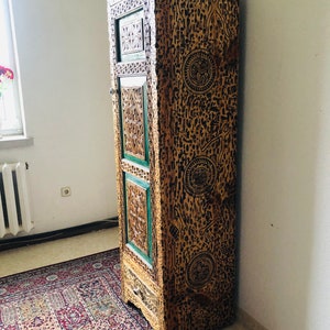 K&ouml;nnte beinhalten: Ein hoher, kunstvoll geschnitzter Holzschrank mit einer gr&uuml;n-braunen Oberfl&auml;che. Der Schrank hat eine einzelne T&uuml;r und eine Schublade. Die T&uuml;r ist mit einem geometrischen Muster verziert.