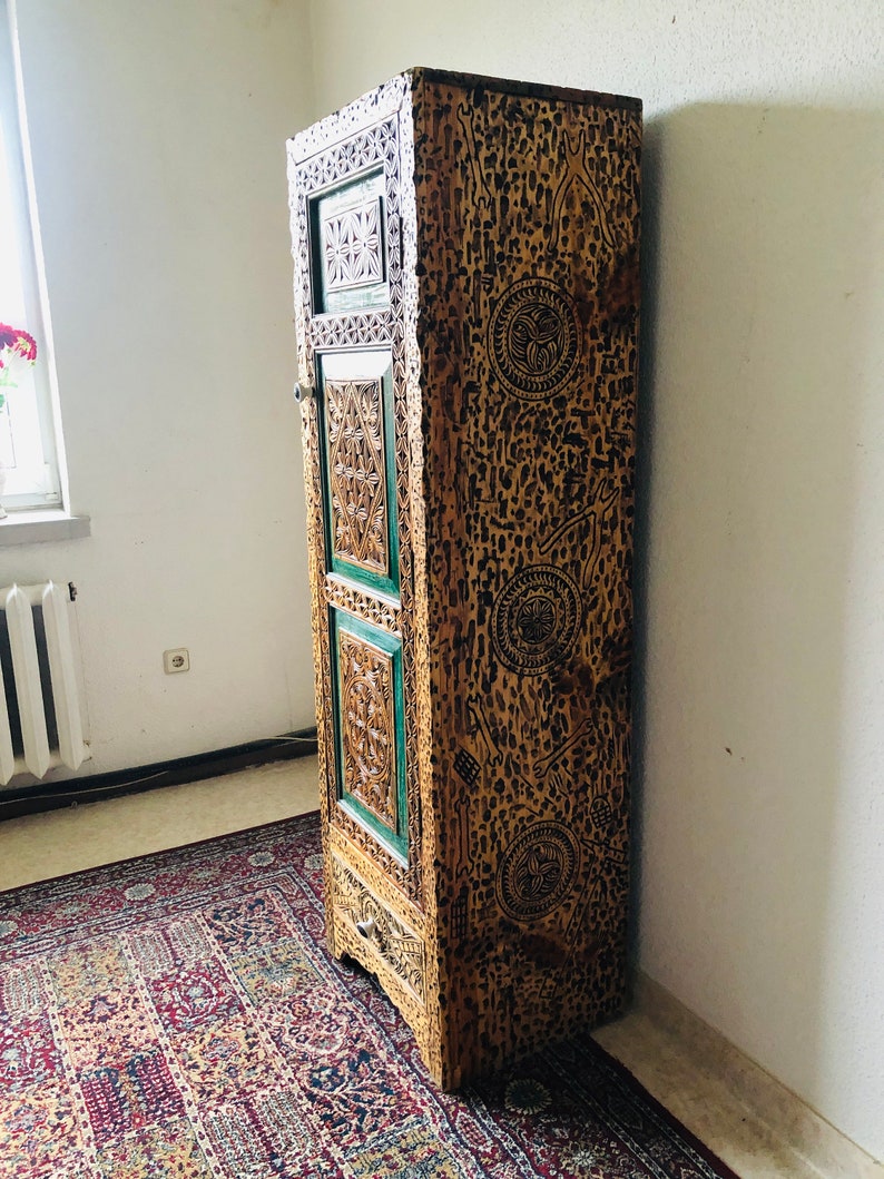 K&ouml;nnte beinhalten: Ein hoher, kunstvoll geschnitzter Holzschrank mit einer gr&uuml;n-braunen Oberfl&auml;che. Der Schrank hat eine einzelne T&uuml;r und eine Schublade. Die T&uuml;r ist mit einem geometrischen Muster verziert.