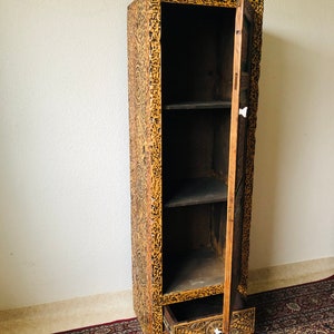 K&ouml;nnte beinhalten: Ein hoher Holzschrank mit einer einzigen T&uuml;r und einer Schublade. Der Schrank ist mit aufwendigen Schnitzereien verziert und hat eine dunkelbraune Oberfl&auml;che.
