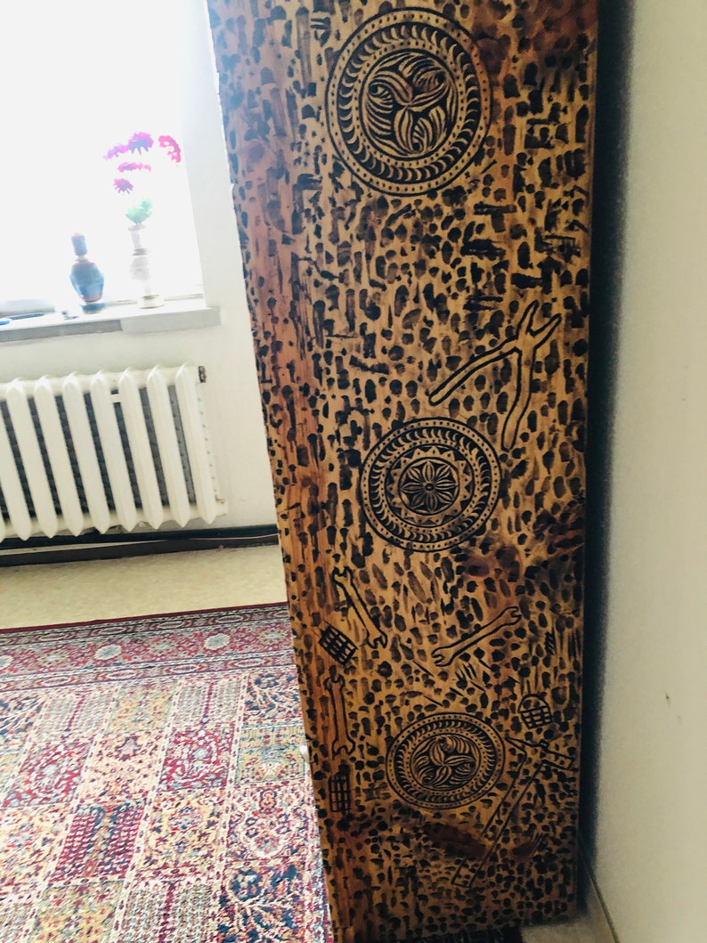 K&ouml;nnte beinhalten: Ein Holzbrett mit brauner Beize und schwarzen Markierungen. Die Markierungen sind kreisf&ouml;rmig und &auml;hneln einer stilisierten Blume. Das Brett ist eine vertikale Fl&auml;che und hat eine raue Textur.