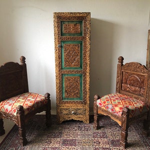 K&ouml;nnte beinhalten: Zwei kunstvoll geschnitzte Holzst&uuml;hle mit bunten gemusterten Kissen stehen vor einem hohen, verzierten Holzschrank mit einer Schublade. Der Schrank ist in Gr&uuml;n und Gold bemalt.