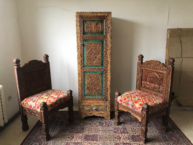 K&ouml;nnte beinhalten: Zwei kunstvoll geschnitzte Holzst&uuml;hle mit bunten gemusterten Kissen stehen vor einem hohen, verzierten Holzschrank mit einer Schublade. Der Schrank ist in Gr&uuml;n und Gold bemalt.