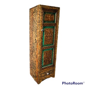 K&ouml;nnte beinhalten: Ein hoher, kunstvoll geschnitzter Holzschrank mit einer einzigen T&uuml;r und einer Schublade. Der Schrank ist in Braun- und Gr&uuml;nt&ouml;nen bemalt, mit kunstvollen floralen und geometrischen Mustern, die in das Holz geschnitzt sind.