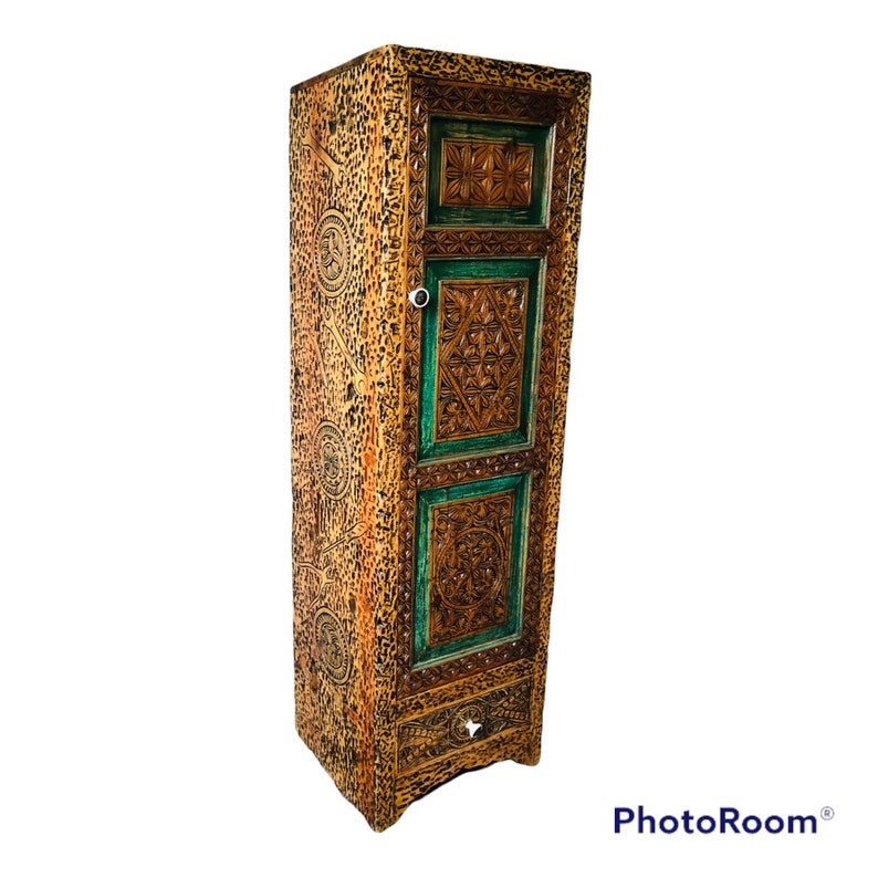 K&ouml;nnte beinhalten: Ein hoher, kunstvoll geschnitzter Holzschrank mit einer einzigen T&uuml;r und einer Schublade. Der Schrank ist in Braun- und Gr&uuml;nt&ouml;nen bemalt, mit kunstvollen floralen und geometrischen Mustern, die in das Holz geschnitzt sind.