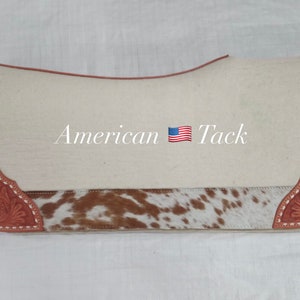Könnte beinhalten: Ein weißer Sattelpad mit braunem und weißem Kuhfellbesatz und braunen Lederakzenten. Der Sattelpad trägt den Text "American Tack" und eine amerikanische Flagge.