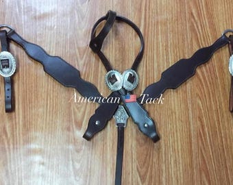 Puede incluir: Bridón de cuero marrón para caballo con conchos plateados y un patrón de costura decorativo. El bridón está colocado plano sobre una superficie de madera. El texto "American Tack" es visible en la imagen.