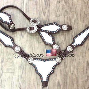 Könnte beinhalten: Ein weißes Leder-Pferdezaumzeug-Set mit braunen Lederriemen und silbernen Conchos. Das Zaumzeug hat einen blauen und roten Akzent an den Rändern. Das Set beinhaltet ein Genickstück, einen Brustlatz und Zügel.