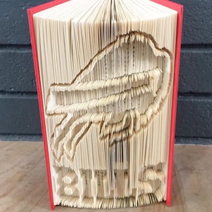 以下が含まれることがあります： ページを折りたたんで鳥の3D彫刻を作った赤い本。彫刻は本のページから作られており、折りたたんで重ねて鳥の形を作っています。