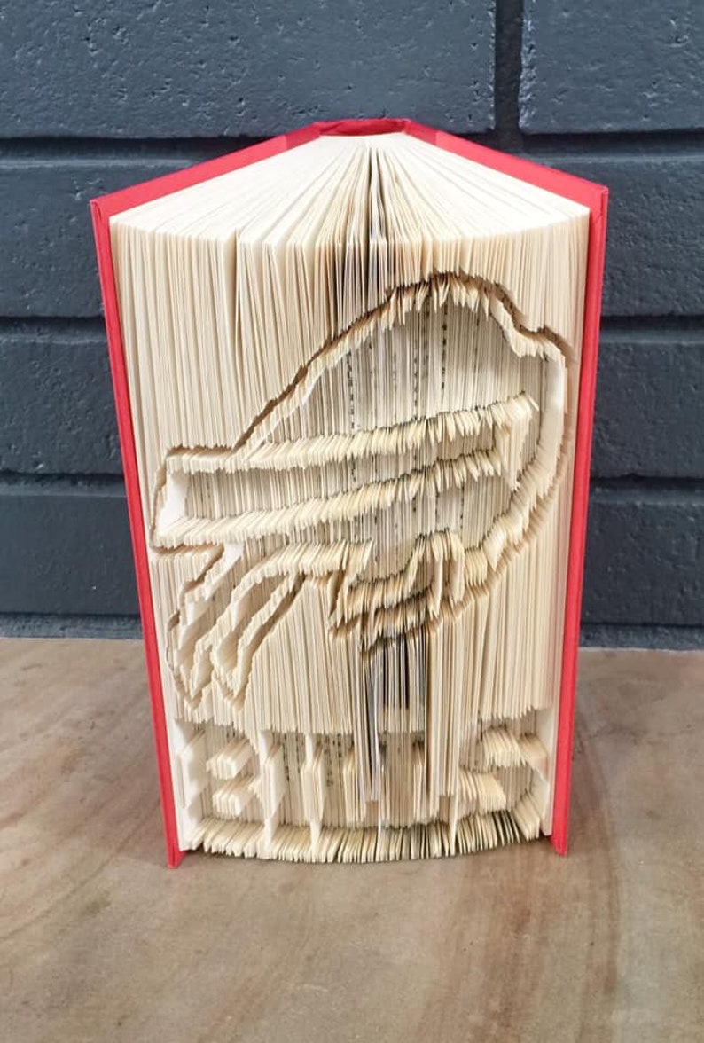 以下が含まれることがあります： ページを折りたたんで鳥の3D彫刻を作った赤い本。彫刻は本のページから作られており、折りたたんで重ねて鳥の形を作っています。