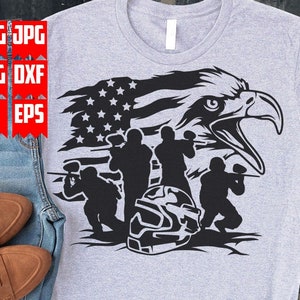 May include: A black and white graphic design featuring a bald eagle with its wings spread out behind it. The eagle is holding a paintball gun in its talons. There are four people in the background, also holding paintball guns. The American flag is behind the eagle and people. The graphic design is on a gray t-shirt.