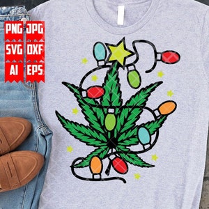 May include: A grey t-shirt featuring a green marijuana leaf design with colourful Christmas lights wrapped around it. The leaf has a yellow star on top.