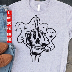 May include: A grey t-shirt featuring a black graphic of a skull giving the middle finger and smoking a joint. The skull is surrounded by smoke and cannabis leaves.