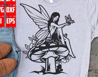 Puede incluir: Un diseño gráfico en blanco y negro de una hada sentada en un hongo con una mariposa en la mano. El hada tiene el pelo largo y alas. El hongo tiene una tapa grande y un tallo. El diseño está en una camiseta gris.