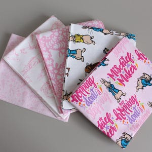 May include: Five square fabric napkins with different patterns. The designs include pink polka dots, white silhouettes on pink, and illustrations of a rabbit in a blue jacket. One napkin has the text "Mischief Maker".