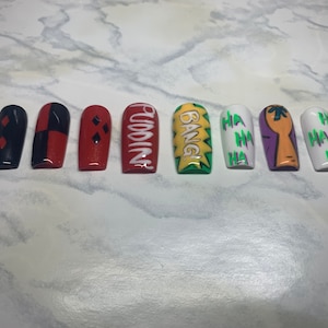 Pode incluir: Um conjunto de dez unhas postiças longas em forma de caixão com vários desenhos. As unhas apresentam uma paleta de cores vermelha, preta, branca, verde e roxa. Algumas unhas têm o texto "Pudding" e "Bang", outras "HA HA".