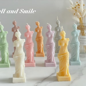 May include: A set of ten colorful, sculpted candles shaped like the Venus de Milo statue. The candles are arranged on a white surface with the text "Smell and Smile" at the top of the image.